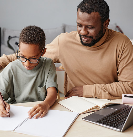 An African American man sits beside a young Black boy while sitting at a table and doing schoolwork.