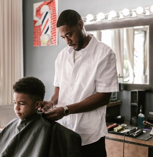 A Black barber adjusts a smock on a young Black boy sitting in a barber chair.